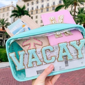 Puede incluir: Bolsa de viaje transparente de plástico con una cremallera turquesa y un "VACAY" turquesa y dorado con purpurina escrito en la parte delantera. La bolsa contiene un portapasaporte rosa con una "M" de conchas en la parte delantera, un portapasaporte beige con un avión de conchas en la parte delantera y una tarjeta de embarque.