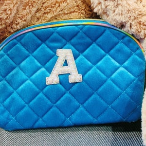 Könnte beinhalten: Eine tealblaue gesteppte Kosmetiktasche mit einem weißen Filzbuchstaben "A", der mit Strasssteinen verziert ist.