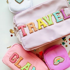 May include: Four pink and purple cosmetic bags with rainbow letters and designs. The bags say "TRAVEL", "GLAM", and have a rainbow and a heart design.
