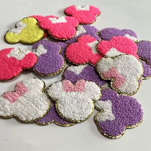 May include: A pile of colorful felt mouse head appliques with gold trim. The appliques are in shades of pink, purple, white, and yellow.