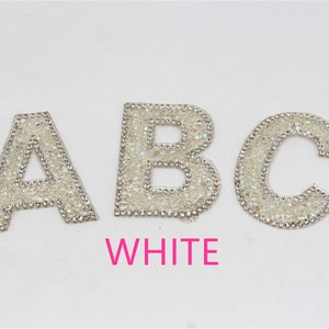 Könnte beinhalten: Drei weiße Strassbuchstaben, A, B und C, auf weißem Hintergrund. Die Buchstaben sind silberfarben umrandet und haben einen glitzernden Finish. Das Wort "WHITE" ist in Pink unter den Buchstaben geschrieben.