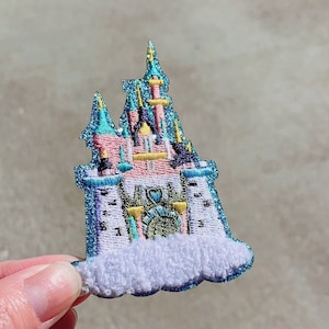 Könnte beinhalten: Ein gestickter Schloss-Patch in Rosa und Blau mit einer weißen, flauschigen Wolkenbasis. Das Schloss hat einen hohen, spitzen Turm und mehrere kleinere Türme. Das Schloss ist von einem blauen und grünen Rand umgeben.