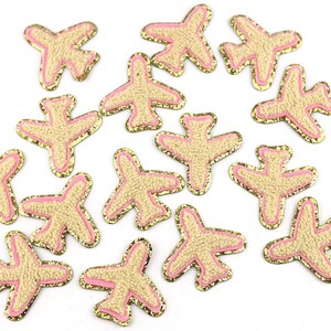 May include: A collection of airplane-shaped patches. Each patch features a textured tan body, outlined with a pink border and a gold glitter edge. The patches are scattered across a white surface.