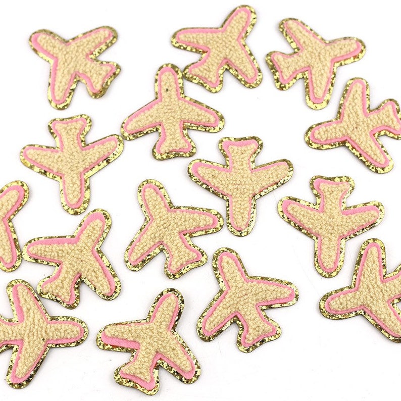 May include: A collection of airplane-shaped patches. Each patch features a textured tan body, outlined with a pink border and a gold glitter edge. The patches are scattered across a white surface.