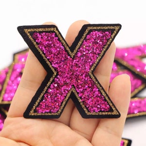 May include: Pink glitter letter X with a black and gold outline. The letter is made of fabric and has a sparkly finish.