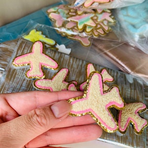 May include: Airplane-shaped patches with a fuzzy, cream-colored body, pink accents, and gold glitter edging. Several patches are visible, some in clear plastic bags. The patches are likely for crafts or embellishments.