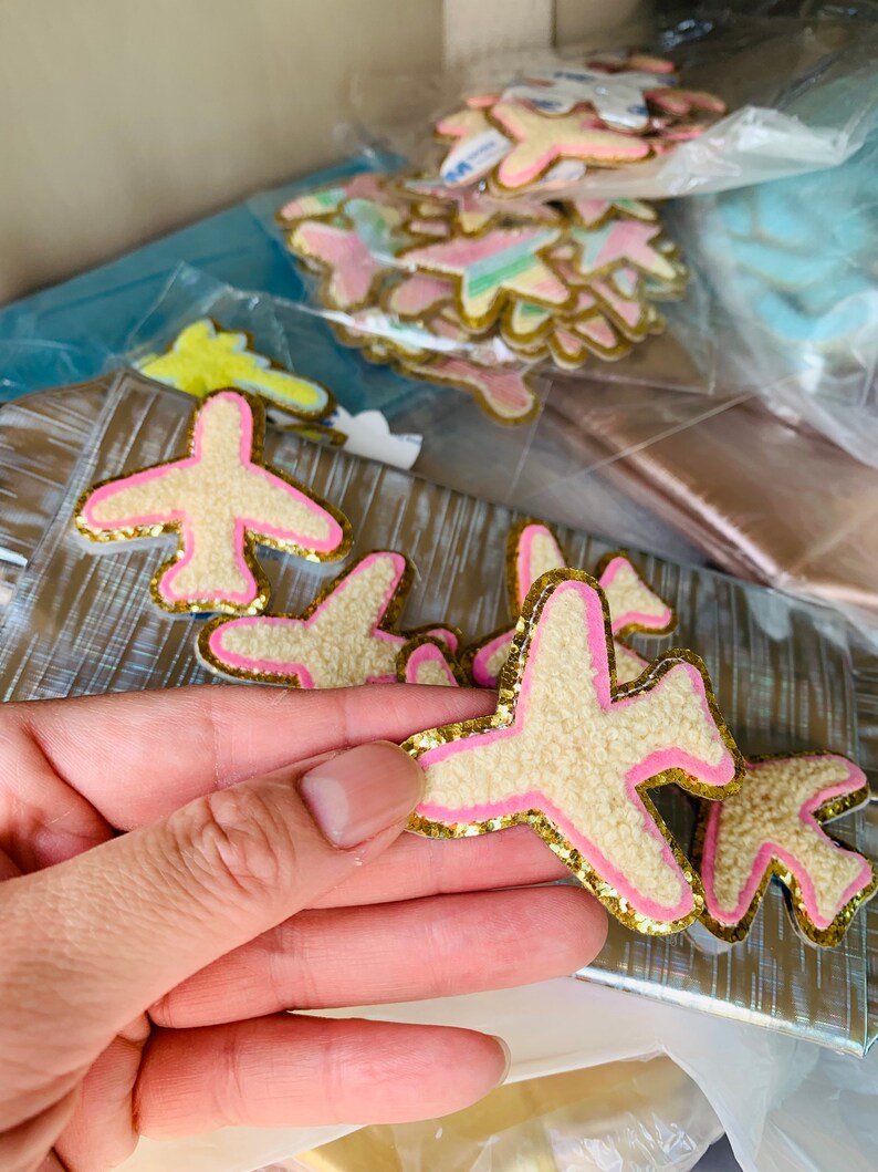 May include: Airplane-shaped patches with a fuzzy, cream-colored body, pink accents, and gold glitter edging. Several patches are visible, some in clear plastic bags. The patches are likely for crafts or embellishments.