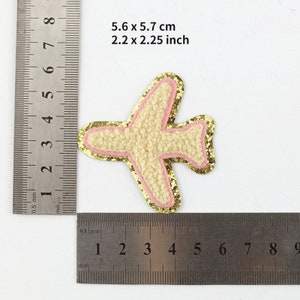 May include: A decorative airplane patch, measuring 2.2 x 2.25 inches. The patch features a textured beige body, outlined with pink and gold glitter. The image includes measurements in both inches and centimeters.