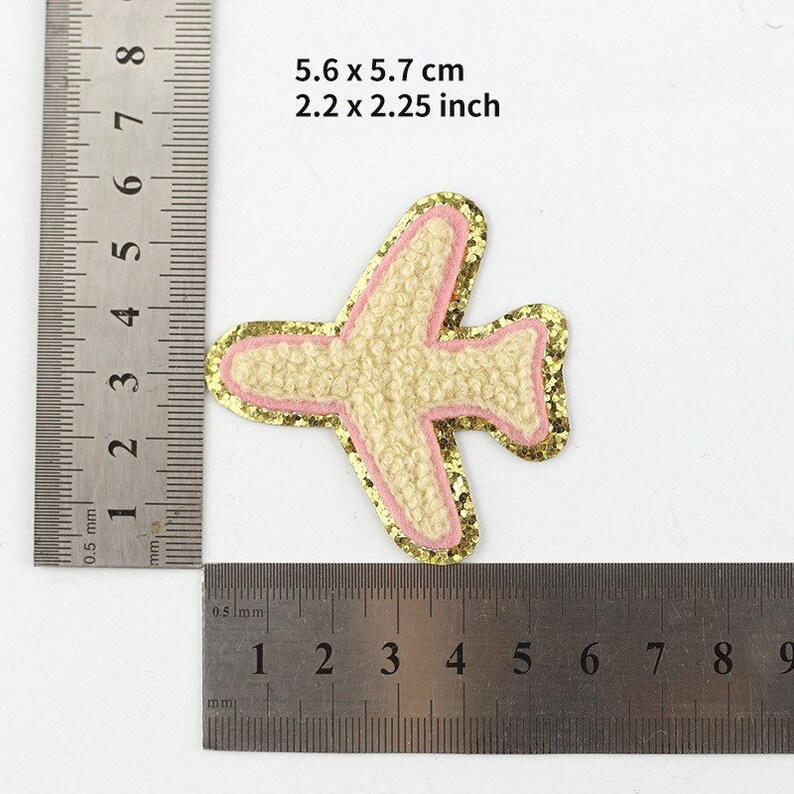 May include: A decorative airplane patch, measuring 2.2 x 2.25 inches. The patch features a textured beige body, outlined with pink and gold glitter. The image includes measurements in both inches and centimeters.