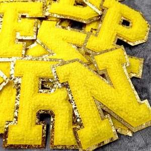 May include: A pile of yellow felt letter patches with gold glitter trim. The letters are all uppercase and are in a variety of sizes. The letters are all in focus and the background is blurred.