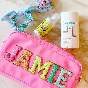 Puede incluir: Una bolsa rosa con cremallera con el nombre "JAMIE" escrito en letras de colores borrosas. La bolsa está al lado de una botella de desinfectante de manos y un stick de desodorante, ambos con la marca "megababe".