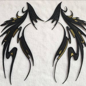 Può includere: Un paio di ali ricamate nere e dorate con disegni intricati. Le ali sono simmetriche e hanno una forma appuntita.