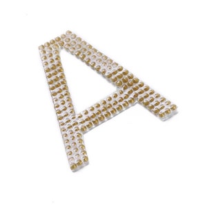 Peut inclure: Une lettre A dorée en forme de petites perles rondes brillantes. La lettre est en relief et a un dos en plastique transparent.