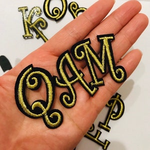 May include: Gold embroidered letters spelling out the name "QAM" on a black background. The letters are outlined in black and have a decorative swirl design.