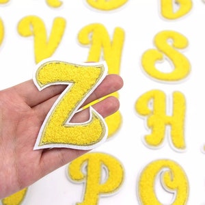 May include: Yellow felt letter patches with a white outline. The letter Z is being held in a hand. The letters are in a cursive style.