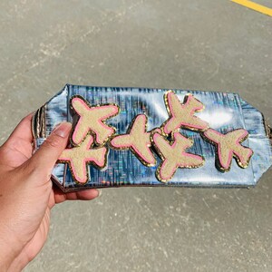 May include: A rectangular clutch with a holographic silver finish. The clutch is decorated with six beige airplane patches with pink and gold trim. The clutch is held by a hand.