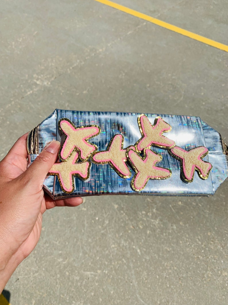 May include: A rectangular clutch with a holographic silver finish. The clutch is decorated with six beige airplane patches with pink and gold trim. The clutch is held by a hand.