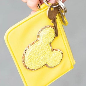 May include: A yellow zippered pouch with a gold glitter Mickey Mouse head design. The pouch is attached to a gold key ring with keys.