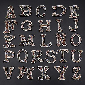 Può includere: Un set di 26 lettere dell'alfabeto da stirare, ogni lettera è decorata con strass e perline colorate. Le lettere sono in uno stile di font classico.