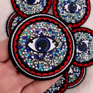 Puede incluir: Un parche redondo bordado en negro, rojo y blanco con un ojo azul rodeado de pedrería transparente, azul y verde. El ojo tiene un iris blanco y una pupilla negra.