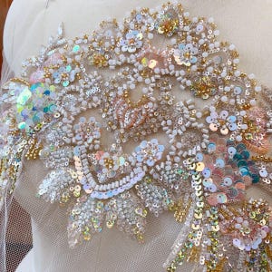 Può includere: Un delicato merletto bianco e dorato con paillettes iridescenti e perline madreperlacee. Il merletto presenta un motivo floreale con dettagli intricati.