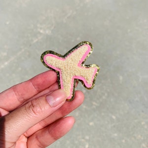 May include: A beige airplane-shaped patch with a pink outline and gold glitter border. The patch has a textured, fuzzy appearance. The design is a fun, travel-themed accessory.