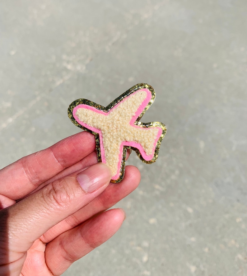 May include: A beige airplane-shaped patch with a pink outline and gold glitter border. The patch has a textured, fuzzy appearance. The design is a fun, travel-themed accessory.