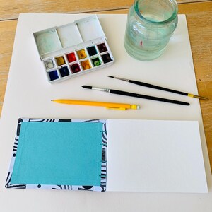 May include: A white watercolor paint set with twelve pans, a clear glass jar filled with water, two black paintbrushes, a yellow mechanical pencil, and an open sketchbook with a black and white patterned cover and a bright turquoise inner page.