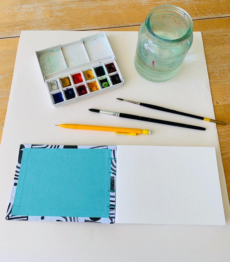 May include: A white watercolor paint set with twelve pans, a clear glass jar filled with water, two black paintbrushes, a yellow mechanical pencil, and an open sketchbook with a black and white patterned cover and a bright turquoise inner page.
