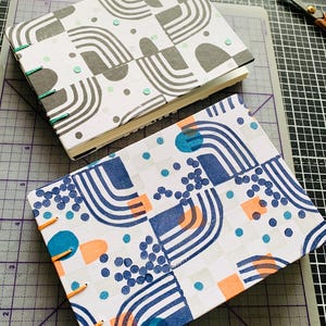 May include: Two rectangular hand-bound journals with exposed stitching along the spine. The top journal features a gray and white geometric pattern with teal accents. The bottom journal displays a blue, orange, and teal geometric print.