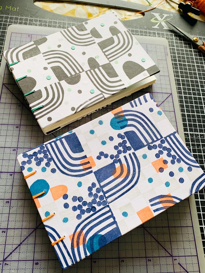 May include: Two rectangular hand-bound journals with exposed stitching along the spine. The top journal features a gray and white geometric pattern with teal accents. The bottom journal displays a blue, orange, and teal geometric print.