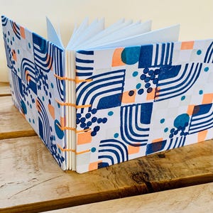 May include: A square notebook with an exposed orange thread binding and white pages. The hard cover features a repeating pattern of dark blue curved lines, solid teal circles, small blue dots, and light orange squares on a white background.