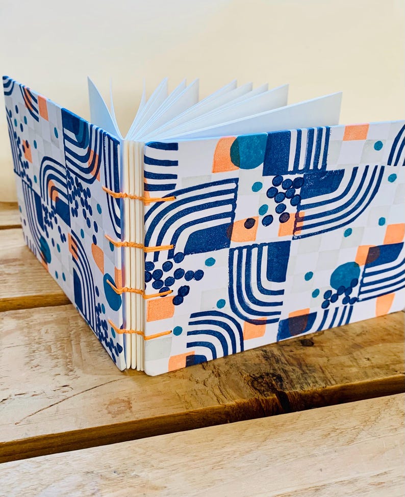 May include: A square notebook with an exposed orange thread binding and white pages. The hard cover features a repeating pattern of dark blue curved lines, solid teal circles, small blue dots, and light orange squares on a white background.