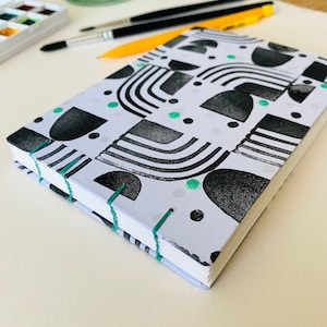 May include: A rectangular notebook with a white cover featuring a black geometric pattern of arches, semicircles, and dots, accented with small green circles. The spine is exposed and bound with bright green thread, showing white paper pages.