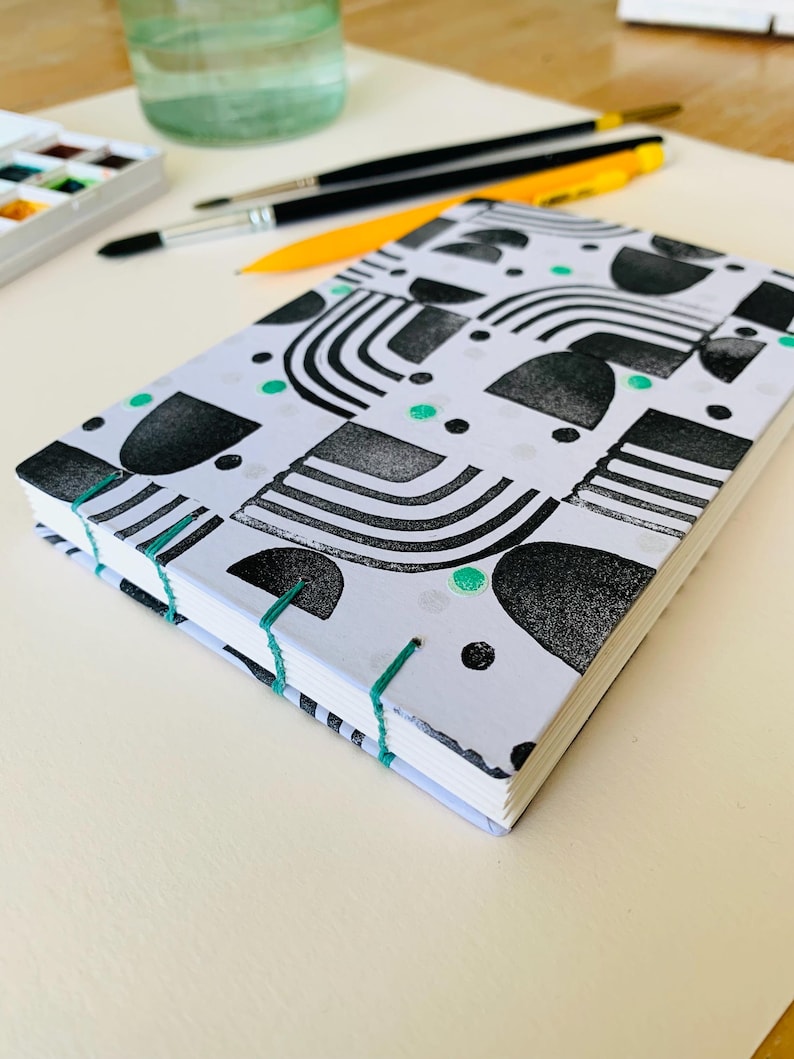 May include: A rectangular notebook with a white cover featuring a black geometric pattern of arches, semicircles, and dots, accented with small green circles. The spine is exposed and bound with bright green thread, showing white paper pages.