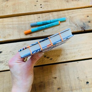 May include: A small notebook with a blue and white patterned cover, featuring exposed orange thread stitching along the spine. Two turquoise and orange fine-tip markers are positioned above the notebook, which has white pages visible at the edge.