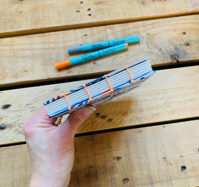 May include: A small notebook with a blue and white patterned cover, featuring exposed orange thread stitching along the spine. Two turquoise and orange fine-tip markers are positioned above the notebook, which has white pages visible at the edge.