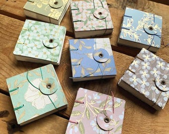 May include: Seven square notebooks with floral patterned covers in various colors including green, blue, pink, and grey. Each notebook features a circular paper closure secured with a metal eyelet and a thin colored string binding the pages together.