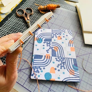 May include: A partially assembled book with a white cover featuring a blue, orange, and gray geometric pattern. The spine is exposed, showing white paper signatures stitched together with bright orange thread, with a spool of matching thread nearby.