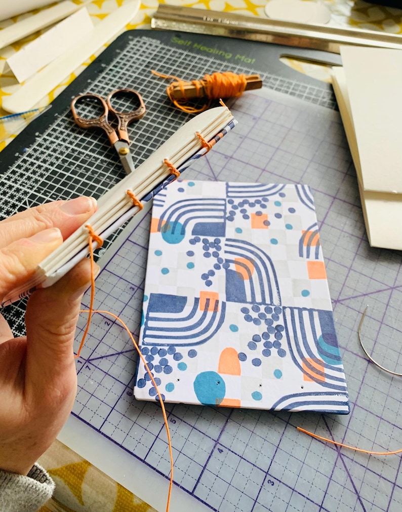 May include: A partially assembled book with a white cover featuring a blue, orange, and gray geometric pattern. The spine is exposed, showing white paper signatures stitched together with bright orange thread, with a spool of matching thread nearby.