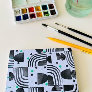 May include: A rectangular notebook with a white cover featuring a black geometric pattern of semicircles, lines, and dots, accented with small teal circles and teal thread binding. Nearby are a watercolor paint set, two brushes, and a yellow pencil.