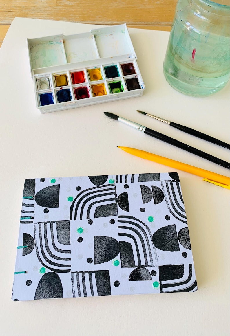 May include: A rectangular notebook with a white cover featuring a black geometric pattern of semicircles, lines, and dots, accented with small teal circles and teal thread binding. Nearby are a watercolor paint set, two brushes, and a yellow pencil.