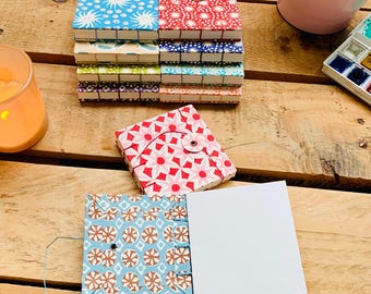 May include: A stack of small, patterned notebooks with button closures. The covers feature various designs in blue, red, green, and purple. One open notebook displays blank white pages. A candle, paintbrushes, and watercolors are also visible.