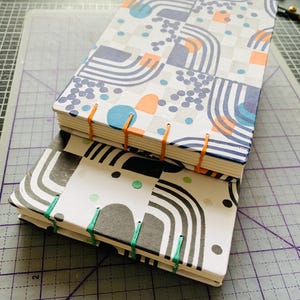 May include: Two hand-bound notebooks with exposed stitching. The top notebook features a white cover with blue, orange, and gray geometric patterns and orange thread. The bottom notebook has a white cover with black geometric shapes and green thread.