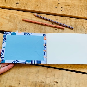 May include: An open notebook with a patterned cover featuring blue and white geometric shapes and a solid light blue rectangular center. The binding is stitched with bright orange thread, and two colored pencils lie above the notebook.