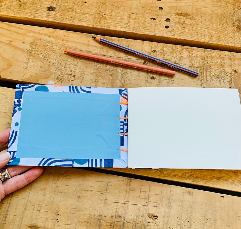 May include: An open notebook with a patterned cover featuring blue and white geometric shapes and a solid light blue rectangular center. The binding is stitched with bright orange thread, and two colored pencils lie above the notebook.