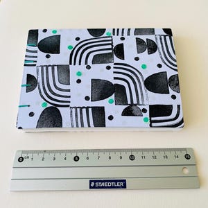 May include: A rectangular notebook with a white cover featuring a black geometric pattern of arches and semicircles, accented with small teal dots. The binding is stitched with teal thread. A 15 cm Staedtler ruler is placed below the notebook.