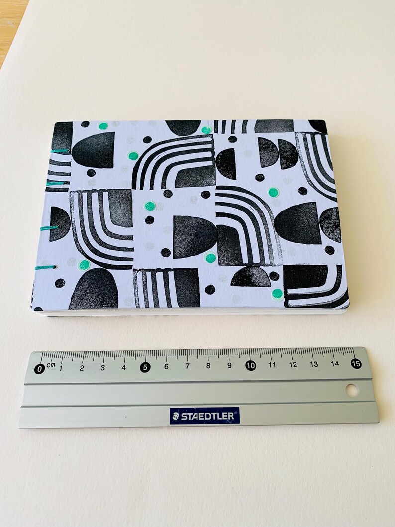 May include: A rectangular notebook with a white cover featuring a black geometric pattern of arches and semicircles, accented with small teal dots. The binding is stitched with teal thread. A 15 cm Staedtler ruler is placed below the notebook.