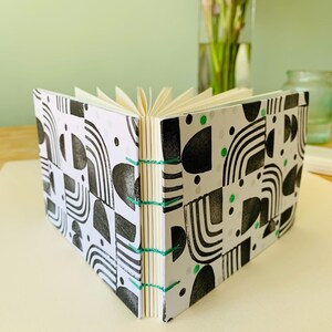 May include: A small, open notebook featuring a white cover with a black geometric pattern of arches and semicircles, accented by small green dots. The pages are bound together with exposed green thread stitching along the central spine.