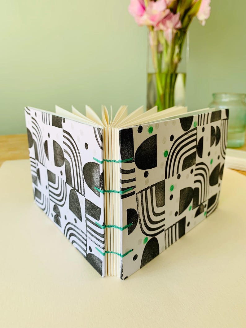 May include: A small, open notebook featuring a white cover with a black geometric pattern of arches and semicircles, accented by small green dots. The pages are bound together with exposed green thread stitching along the central spine.